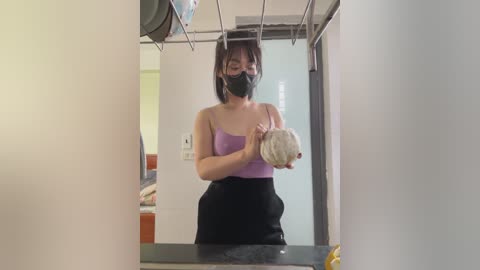 Media: Video of an Asian woman with short black hair, wearing a black mask, purple tank top, and black skirt, holding a fluffy white cat in a modern, clean bathroom.