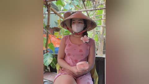 Media: Video of a young Asian woman with light skin, wearing a pink floral dress, conical hat, and face mask, sitting in a green, rustic, outdoor garden setting.