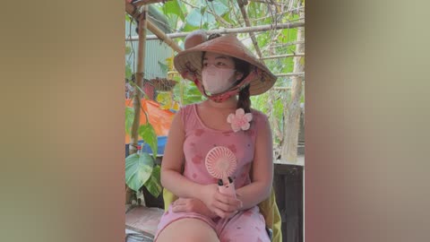 Media: Video of a young Southeast Asian woman with fair skin, wearing a pink dress, straw hat, and face mask, seated on a wooden bench amidst lush greenery and colorful fabric decorations.