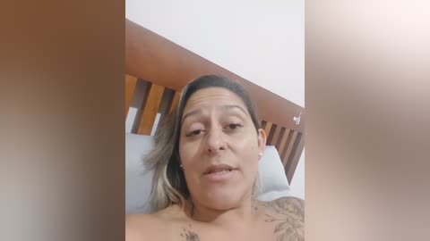 Media: Video of a Latina woman with light brown skin, medium build, and medium-length hair, lying on a bed with a wooden headboard. She has a tattoo on her right shoulder and is topless.