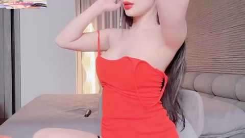 Video of a fair-skinned woman with long, straight, dark hair, wearing a tight, bright red dress with thin straps, posing seductively with her arms raised. Background includes a bed with a beige headboard, a beige lamp, and a blurred TV screen.