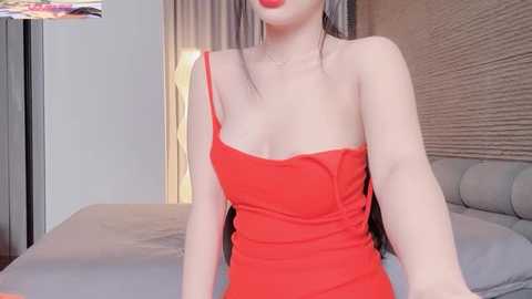 Video of an Asian woman with fair skin, wearing a tight, red, strapless dress, sitting on a bed in a modern, beige-toned bedroom.