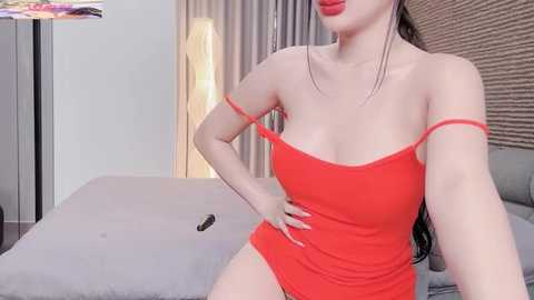 Video of a woman with light skin, wearing a tight, bright red, strapless dress, sitting on a bed with a grey blanket. Her long black hair is pulled back, and she has a confident pose. The room has beige curtains and a lamp in the background.