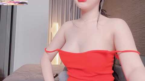 Video of a fair-skinned woman with long, dark hair wearing a strapless red dress, showcasing her cleavage, standing indoors near a bed.