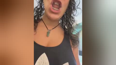 Media: Video of a young woman with curly dark hair, wearing a black tank top with white abstract designs, and a green pendant necklace. She has her mouth open, showing braces. Background is blurred.