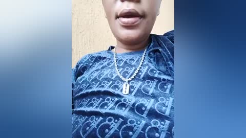 Media: Video of a person with medium brown skin, wearing a blue denim shirt with a geometric pattern, a silver necklace with a pendant, and a closed-mouth smile. Background is a gradient of blue and beige.