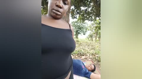 Media: Video of a plus-size Black woman with short hair, wearing a black tank top, standing in a lush, green, outdoor setting with a man lying on the ground in the background.