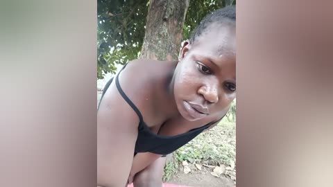 Media: Video of a young Black woman with short hair, leaning forward, wearing a black tank top, standing outdoors near a tree with green foliage and grass.