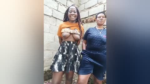 Media: Video of two African women standing against a white-brick wall. The woman on the left, with medium-dark skin and long braids, wears an orange crop top exposing her breasts and a black and white patterned skirt. The woman on the right, with medium skin tone and short hair, wears a navy blue dress.