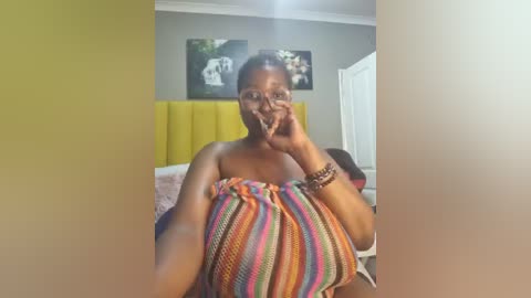 Media: Video of a smiling Black woman with dark skin, wearing a colorful, striped blanket, standing in a modern bedroom with yellow headboard and framed art on the wall.