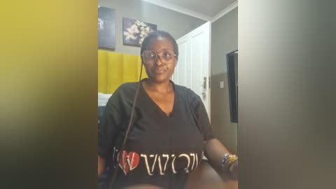 Media: Video of a Black woman with short braided hair, wearing glasses, a black t-shirt, and a crossbody bag, standing in a modern room with gray walls, a yellow blanket, and a TV.