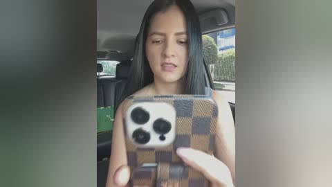 Media: A video of a young woman with long, straight black hair, light skin, and a neutral expression, taking a selfie inside a car. She's wearing a brown and beige checkered phone case.