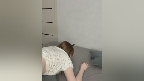 Media: Video of a person with short brown hair, lying on a gray bed with a white nightgown, in a small, plain room with white walls and a closed closet.