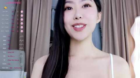 Media: A video of an East Asian woman with long black hair, fair skin, and a slender physique, smiling. She wears a white spaghetti-strap top. Background shows beige curtains and a digital screen with chat options.