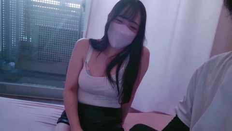 Media: A video of an Asian woman with long black hair, wearing a white mask and a white tank top, kneeling on a bed in a dimly lit room.