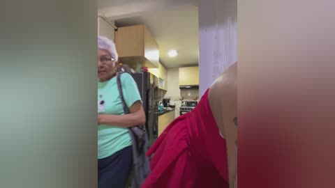 Media: Video of an older woman with short white hair, wearing a green t-shirt, standing in a narrow hallway, looking towards a kitchen with beige walls and a hanging coat.