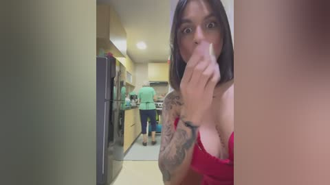 Media: Video of a woman with long dark hair and a tattooed arm, in a red top, holding a phone, standing in a kitchen with a shocked expression.