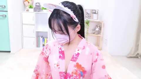 Media: A video of an East Asian woman with long black hair, wearing a pink floral kimono, face mask, and white headband, seated indoors with a white bookshelf and stuffed toys in the background.