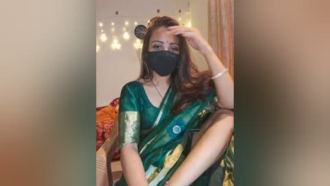 Media: Video of a young woman with long brown hair, wearing a green saree with gold embroidery, black face mask, and silver bracelet. She sits on a bed with a red and orange quilt, surrounded by warm fairy lights in a cozy, softly lit room.