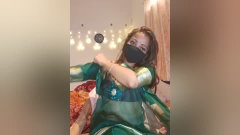 Media: Video of a woman in a green sari with gold embroidery, black face mask, and a light brown complexion, posing in a cozy, softly lit bedroom with string lights and a colorful bedspread.