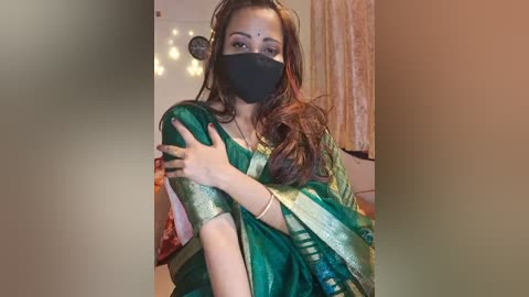 Media: Video of a South Asian woman with light brown skin, long dark hair, wearing a green and gold saree, black mask, and gold bangles. Background features soft lighting and a wooden wall.