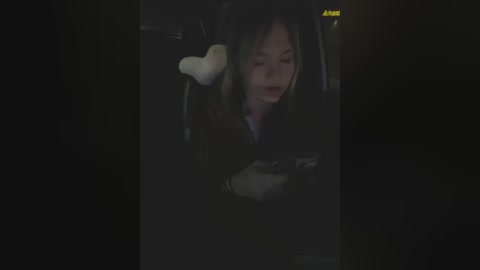 Media: A dimly lit video of a woman with shoulder-length hair, wearing a black coat, sitting in a car with a white teddy bear in her lap, her eyes closed.