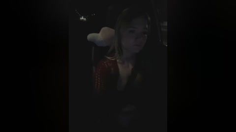 Media: A dimly lit video of a young woman with shoulder-length brown hair wearing a red sequin dress, partially hidden by a fluffy white headpiece, standing in a dark, possibly nighttime, setting.