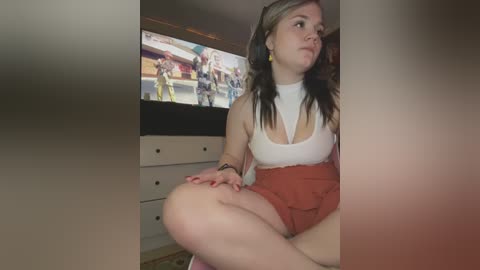 Media: Video of a young woman with light skin, dark hair, and fair complexion, wearing a white top and red shorts, sitting on a bed, watching TV, with a video game character on the screen in the background.