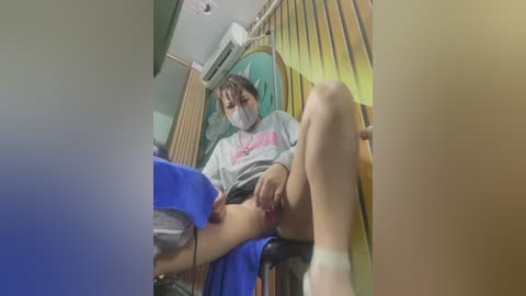 Media: Video of a woman with medium skin tone, wearing a gray hoodie, face mask, and shorts, sitting on a chair with a blue towel, in a room with yellow curtains and a wooden fan.