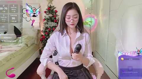 Media: A video of an East Asian woman with glasses and straight black hair, wearing a white blouse, sitting in a bedroom with a Christmas tree and a phone screen.