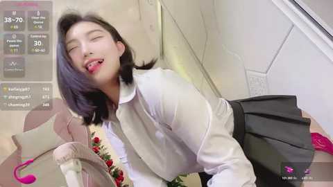 Media: Video of an Asian woman with straight black hair, wearing a white blouse, black skirt, and pink lipstick, lying on her stomach on a pink couch, with a rose necklace. Background shows a white wall with a power outlet.