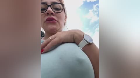 Media: A video of a woman with glasses and fair skin, wearing a light blue top, leaning forward. Her hand with red nail polish rests on her breast, and she has a white wristwatch.