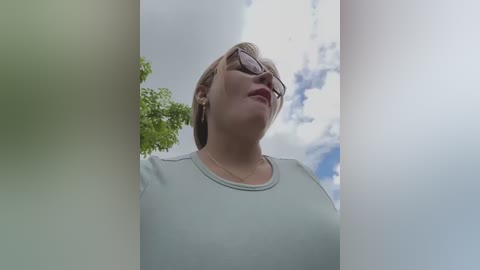 Media: Video of a smiling woman with short brown hair, wearing glasses and a light grey top, standing in a bright, cloudy sky with a green tree in the background.