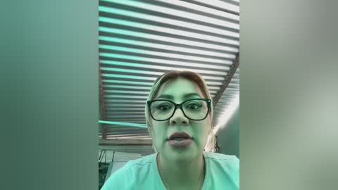 Media: Video of a woman with light skin, short brown hair, and glasses, wearing a white top, standing in a room with a metallic, slatted ceiling and green lighting.