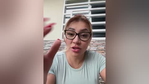 Media: Video of a young Asian woman with fair skin, light brown hair, and glasses, wearing a light blue t-shirt, making a hand gesture, with a stone wall and window in the background.