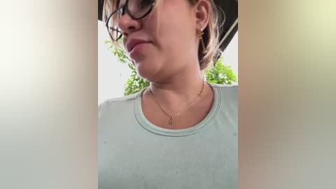 Media: Video of a light-skinned woman with brown hair in glasses, wearing a light grey T-shirt and a delicate gold necklace, outdoors with green foliage in the background.