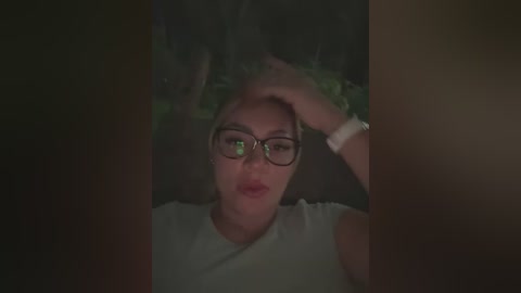 Media: A dimly lit video shows a woman with a pale complexion, wearing black-rimmed glasses, a white top, and a smartwatch. She has a worried expression, hands on her head. The background is dark, with indistinct shapes, suggesting a private, intimate setting.