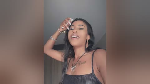 Media: Video of a light-skinned Black woman with long black hair, wearing a black sleeveless top, applying makeup in a dimly lit room with neutral walls.