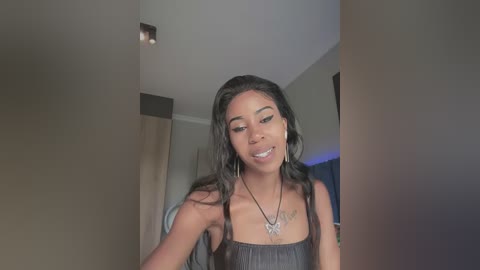 A video of a young Black woman with long, wavy black hair, wearing a black tank top and hoop earrings, smiling slightly while adjusting her hair. She's in a modern, dimly lit room with beige walls and a recessed light fixture.