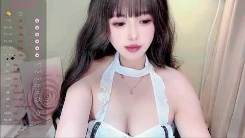 Media: Video of a young Asian woman with long black hair, fair skin, wearing a white halter top that highlights her ample breasts, and a delicate necklace. Background shows a beige curtain and a pink rose patterned cushion.