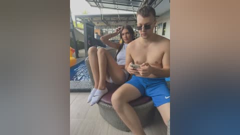 Media: Video of a young woman with long brown hair in a white tank top and shorts sitting on a wicker chair next to a shirtless man in blue shorts, both looking at their phones.