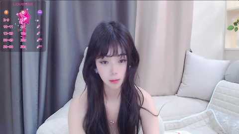 Media: Video of an Asian woman with long black hair, fair skin, and red lipstick, sitting on a white couch in a modern living room with gray curtains.