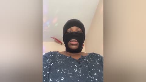 Media: Video of a person with dark skin, wearing a black mask and a dark, patterned top, standing against a blurred, beige background.