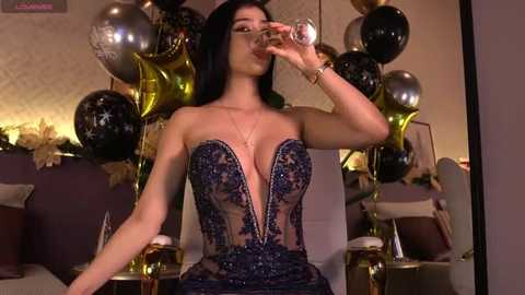 Video of a glamorous woman with long black hair, wearing a revealing, dark blue lace bodysuit, drinking from a glass, surrounded by festive gold and black balloons in a warmly lit room.