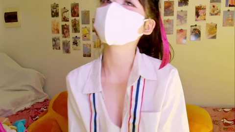 Video of a young woman with light skin, wearing a white lab coat, pink hair, and a face mask, standing in a room with a bed, yellow wall, and a collage of photos.