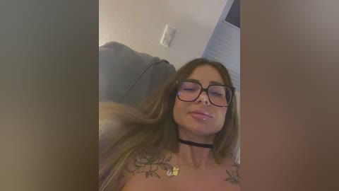 Media: A video of a young woman with long brown hair, glasses, and a black choker, lying on a leather couch. She has tattoos on her shoulders and arms. The background shows a white wall and a light switch.