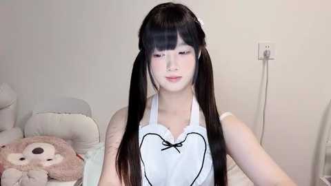 Media: Video of a young Asian woman with long black pigtails, fair skin, and a petite frame, wearing a white apron with a black bow, sitting on a beige couch in a minimalist room.