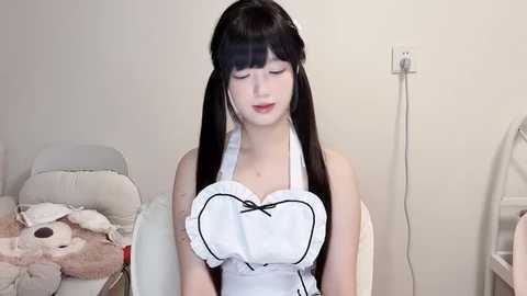 Media: Video of a young, pale-skinned woman with long black hair, wearing a white maid outfit, sitting in a white chair against a beige wall. Background includes a white bed and a power outlet.