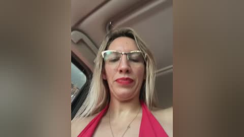 Media: A video of a blonde woman with fair skin, wearing glasses and a red halter top, seated in a car with beige interior, looking down.