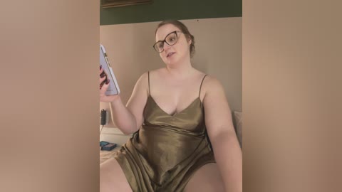 Media: Video of a plus-sized, fair-skinned woman with glasses, wearing a green satin slip dress, holding a blue clipboard. She sits on a beige couch in a beige-walled room, appearing to be taking notes.